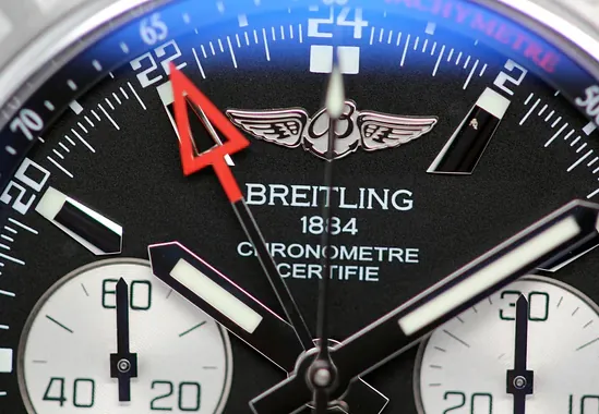 Vedere macro a cadranului unui cronograf Breitling, evidențiind măiestria exterioară premium: logo lustruit, marcaje orare luminescente, text de certificare cronometru imprimat cu precizie și iconica manetă GMT roșie cu finisaj de înaltă calitate.